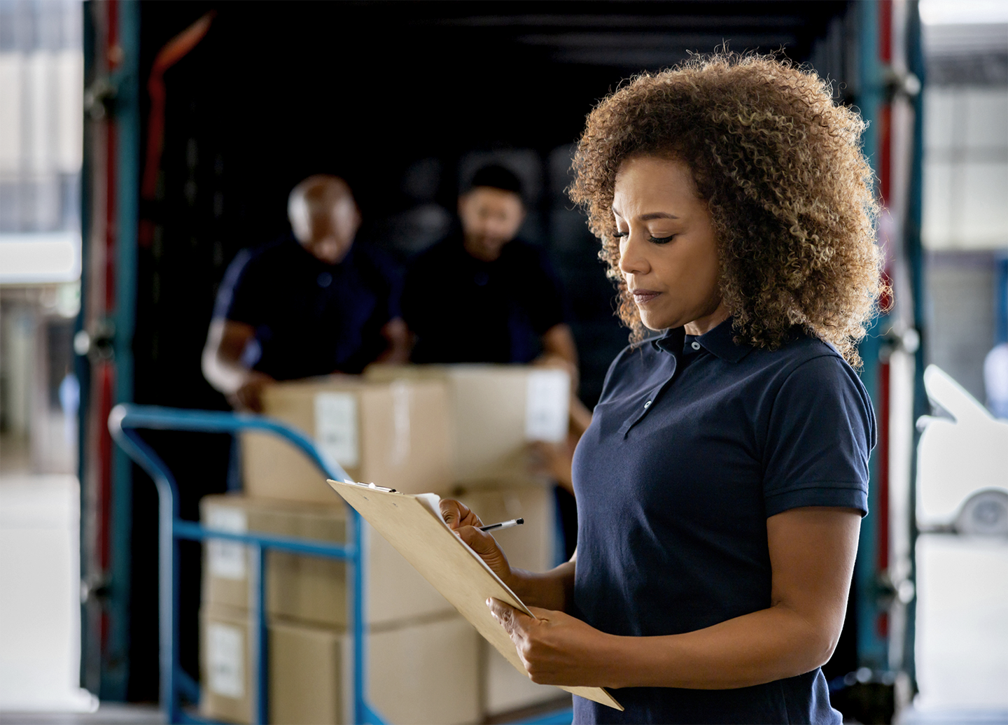 Woman with clipboard inspecting inventory as two workers unload boxes at a loading dock in a warehouse.